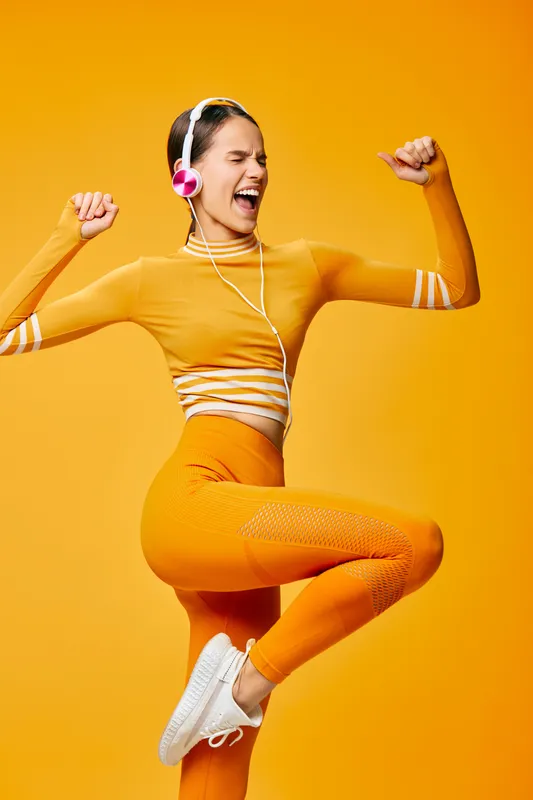 Vibrant woman in orange sportswear, headphones on, mid jump in a bright studio, showcasing fitness, sport and high-energy workout moments.