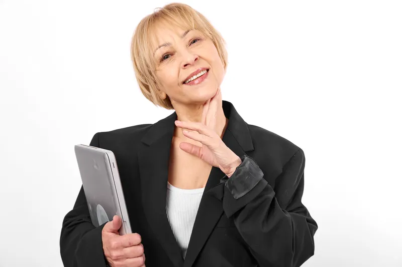 Smiling senior woman holds a smartphone, hand on neck, studio portrait on white background, soft light Concept of communication, mobile technology, wellness, confidence, support