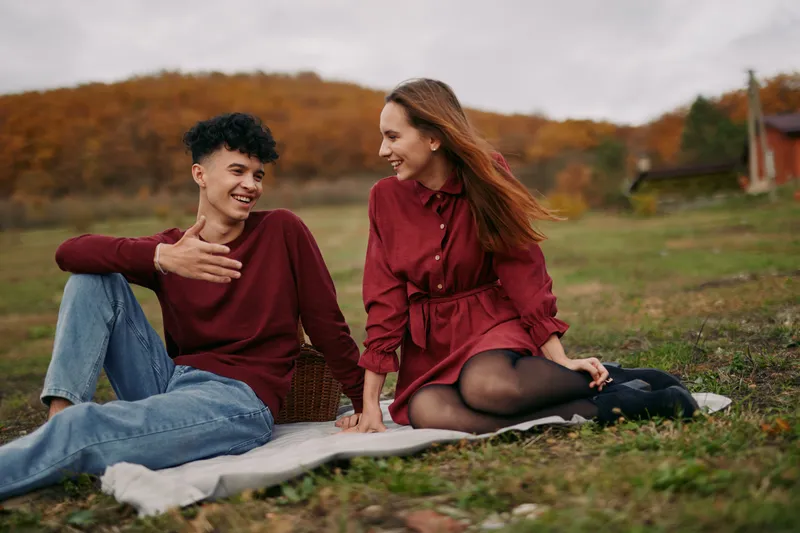 Candid young couple sitting on a blanket in an autumn meadow, sharing warm conversation and laughter, portraying an authentic connection and credible everyday emotion in natural light.