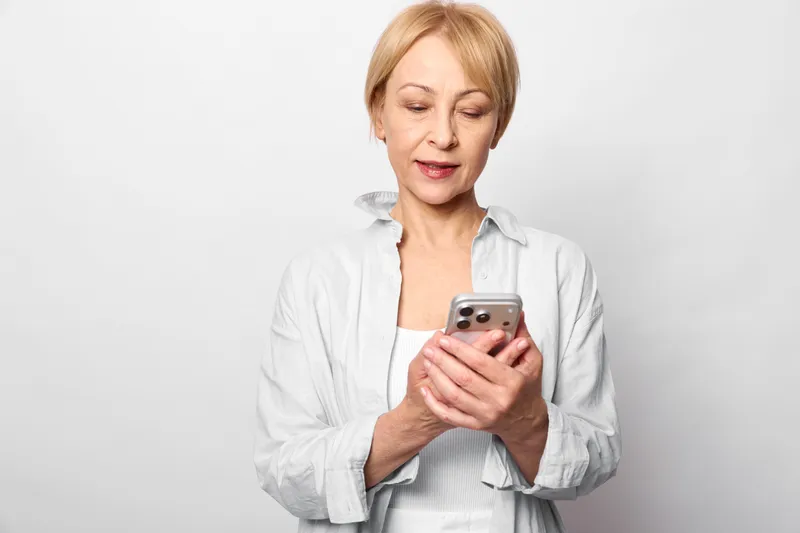 Active mature woman holds smartphone with stress and anxiety, sad tired expression, she reads messages for communication and texting, clean white studio background, concept emotional strain