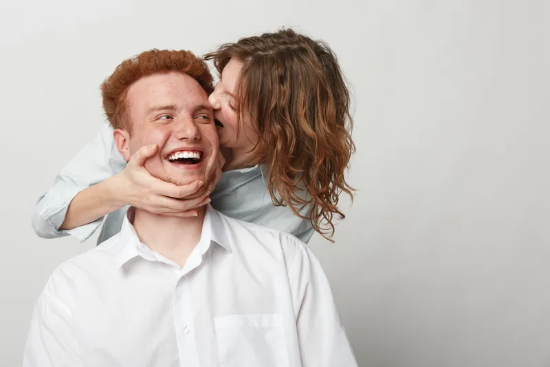 Happy young couple in casual clothes share joyful moment, woman kissing man on cheek. White background, positive emotions, love, affection, and relationship concept.
