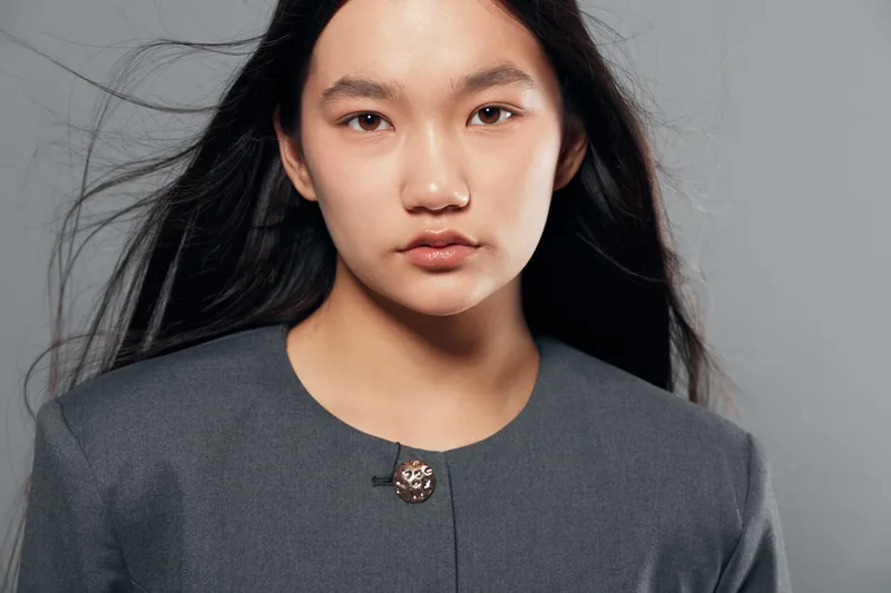 Portrait of confident young woman with long black hair and natural makeup wearing gray formal jacket against neutral background with serious expression and direct eye contact.