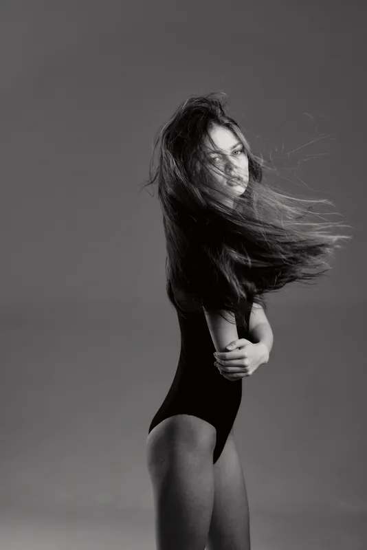 Black and white portrait of a young woman with long hair posing with crossed arms, showing elegance and motion in a simple studio setting.