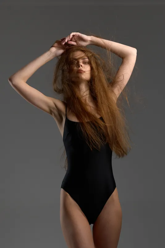 Young woman with long hair in black bodysuit posing with eyes closed against gray background