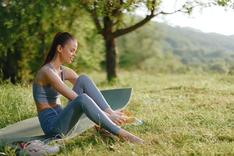 Young fitness woman enjoying healthy snack outdoors, wearing sportswear in a lush green park, representing healthy lifestyle and wellness concepts