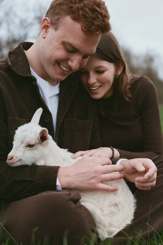 Young couple gently holds a white baby goat outdoors while sharing a tender moment in casual autumn clothing on a cloudy day in a natural setting.