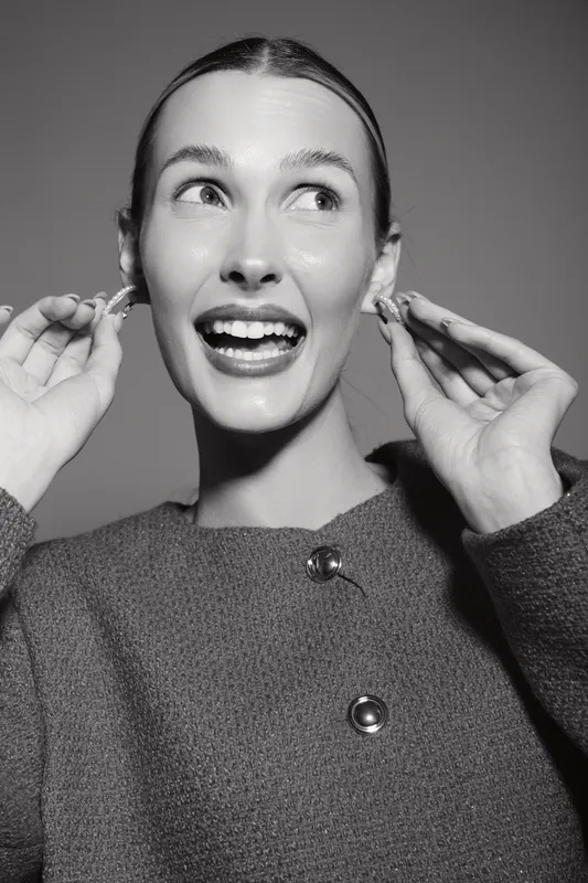 Happy Caucasian woman with fair skin and pulled back hair smiles widely while holding earrings near ears. Studio portrait in black and white. Concept of joy, fashion, and beauty.