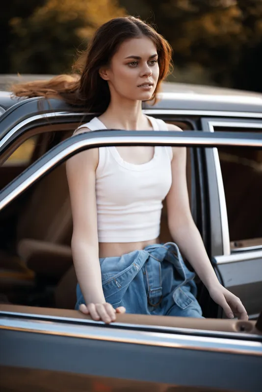 Young woman in casual outfit stepping out of a vintage car, expressing confidence and freedom in a natural outdoor setting during sunset.