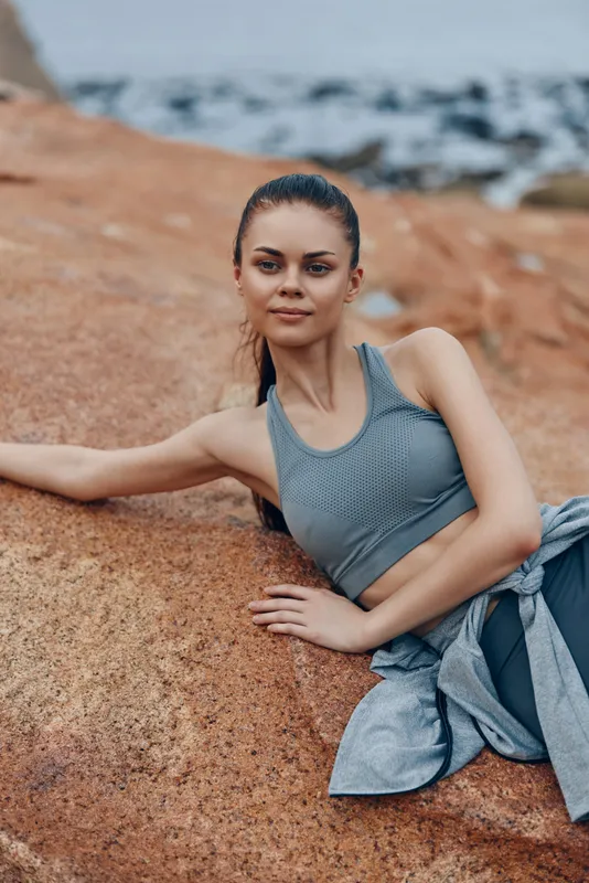 Young woman in athletic wear poses on rocky beach, showcasing a healthy lifestyle with serene emotions, vibrant colors, and a natural background reflecting fitness and nature