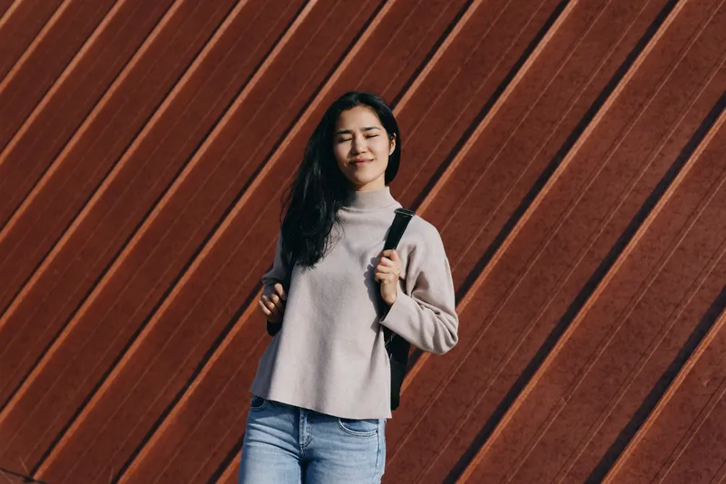 Woman with backpack standing against a diagonal wooden wall in a casual sweater and jeans, urban lifestyle scene showcasing travel style, everyday outfit and confidence