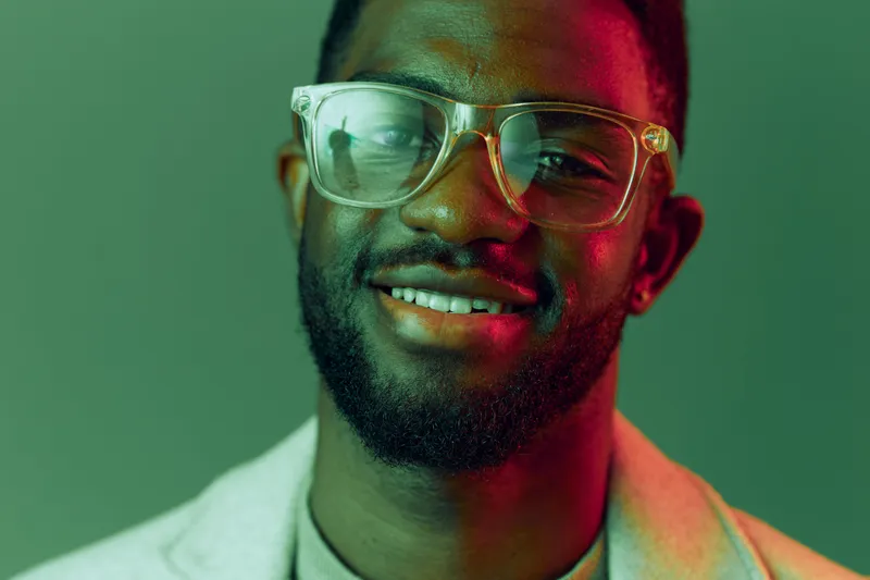 Smiling young African man with glasses in a stylish outfit, exuding positivity against a vibrant green background, perfect for modern lifestyle themes