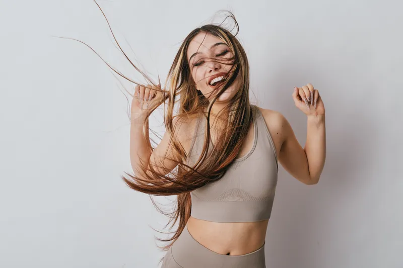 Woman dancing with long hair and smiling in casual top and leggings against light background, enjoying movement and happiness in studio setting with carefree expression.