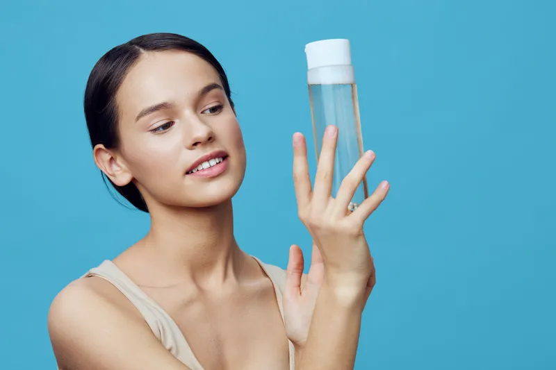 Young woman examining a skincare product in a light blue background, representing healthy lifestyle, beauty routine, and self care practices