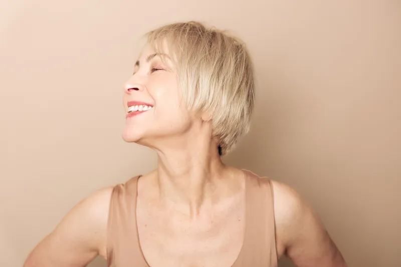 Smiling mature woman with short blonde hair in beige top poses with closed eyes on light background. Natural beauty and happiness concept with soft studio lighting and warm tone.
