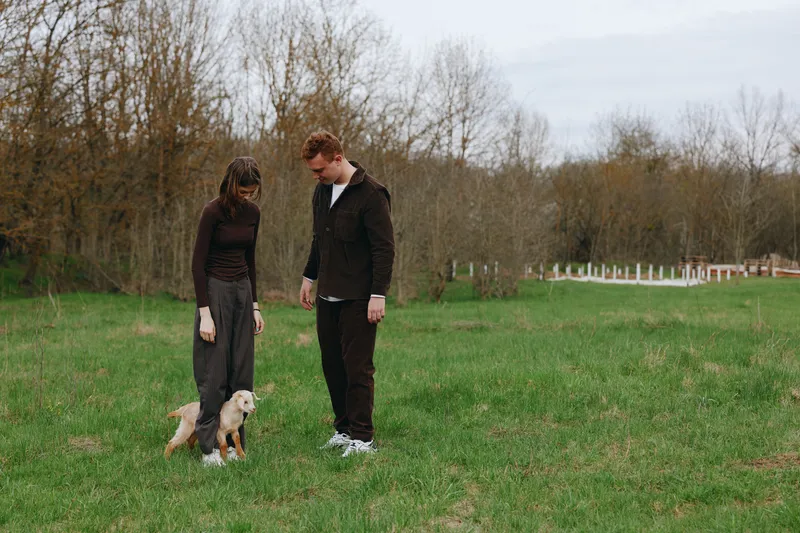 Casual couple walks in a green field with a small dog, enjoying a relaxed outdoor moment, natural light, simple outfits, warm mood, candid lifestyle scene in an open park
