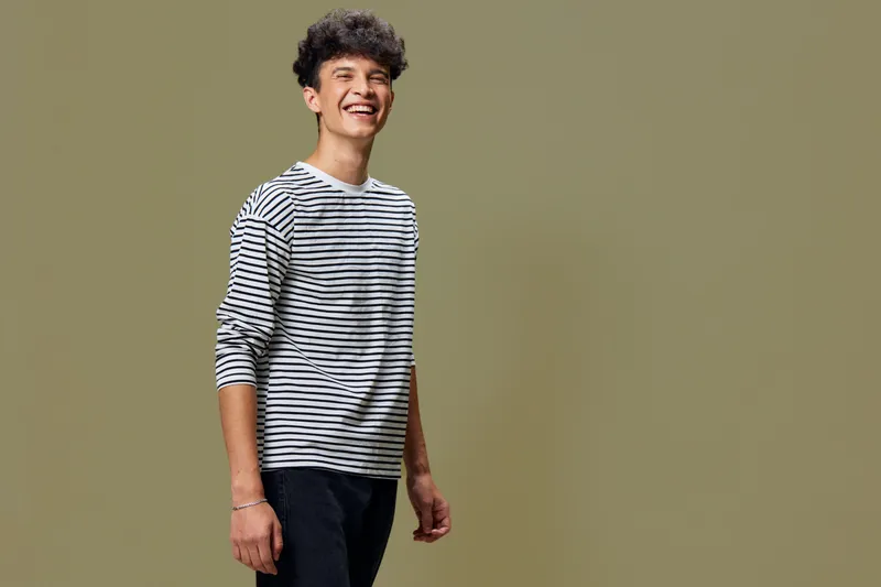 smiling man wearing striped shirt with dark pants stands against beige background with cheerful expression and casual posture in natural lighting