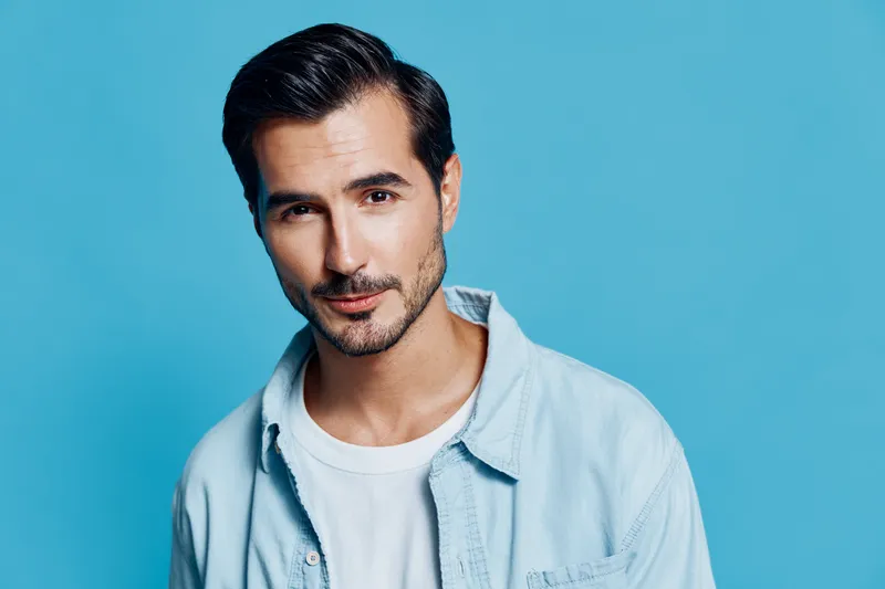 Handsome confident young man wearing a light blue shirt smiles at the camera against a vibrant blue background. People lifestyle concept.