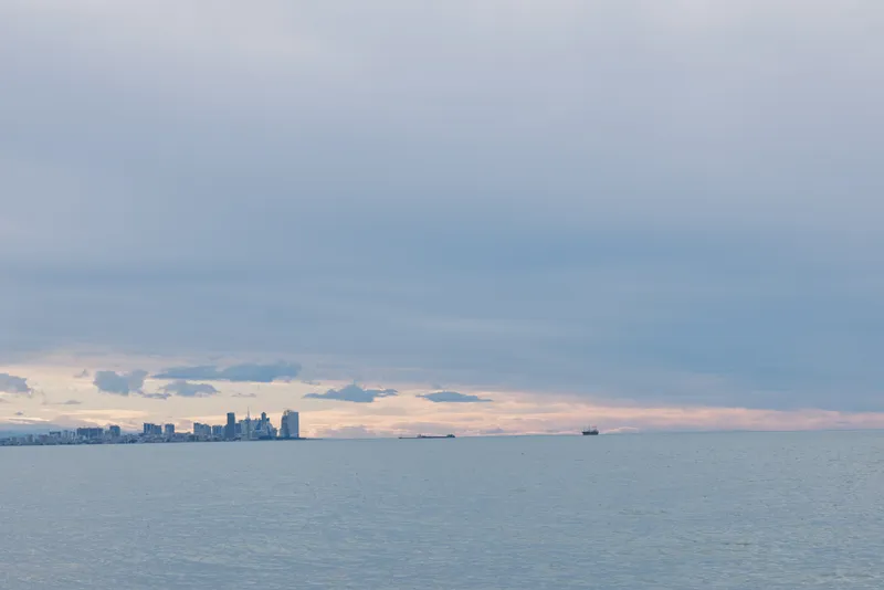 Ocean and distant city skyline on the horizon under calm clouds, boats on the water, serene seascape capturing wide horizon and gentle sunset glow over the sea