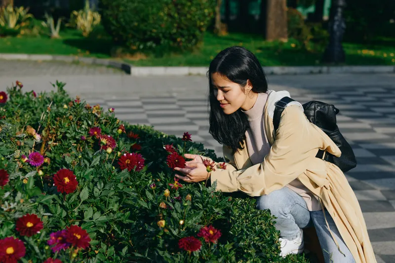 Garden scene with a woman kneeling beside flowering blooms, tending plants in a light coat, outdoors calm and nature inspired, showcasing care, growth and beauty of the garden