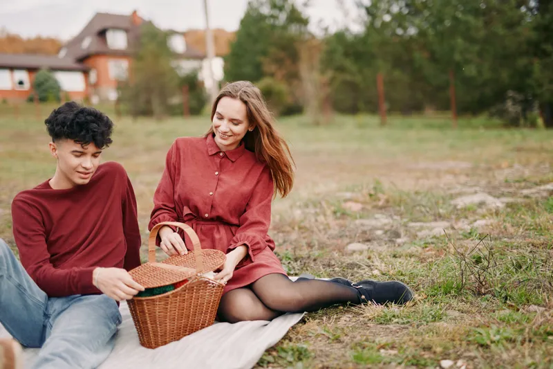 Candid outdoor picnic of a young couple sharing a wicker basket, conveying authenticity and credibility through genuine smiles, natural gestures and relaxed everyday connection in autumn.