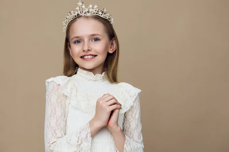 A cheerful young girl wearing a delicate lace dress and a tiara, posing happily against a soft beige background, embodying innocence and joy
