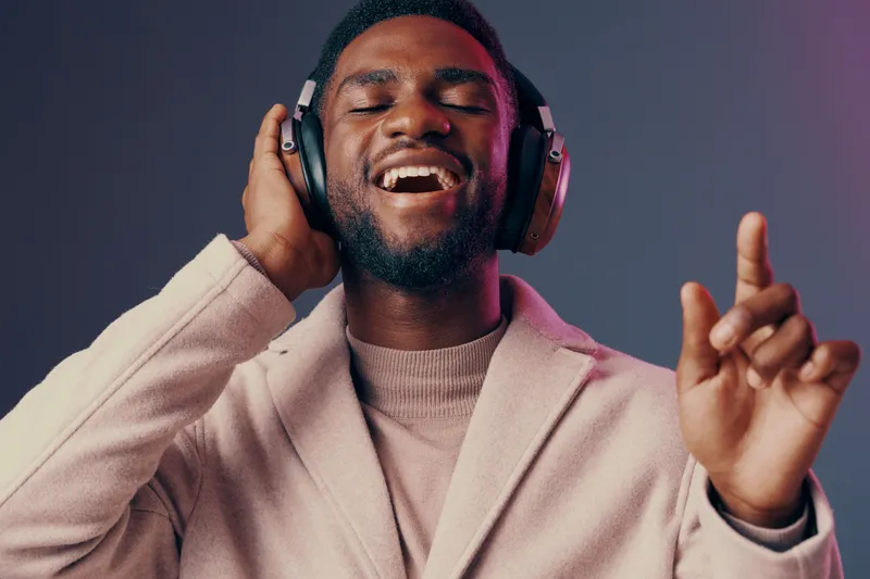 Happy Black man enjoying music with headphones in modern attire, smiling joyfully against a soft gradient background Perfect for wellness and lifestyle themes