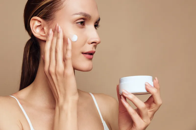 Young woman applying white face cream on cheek with fingers, holding jar in hand. Close-up on beige background. Concept of skincare, beauty, and daily self-care routine with soft natural light.