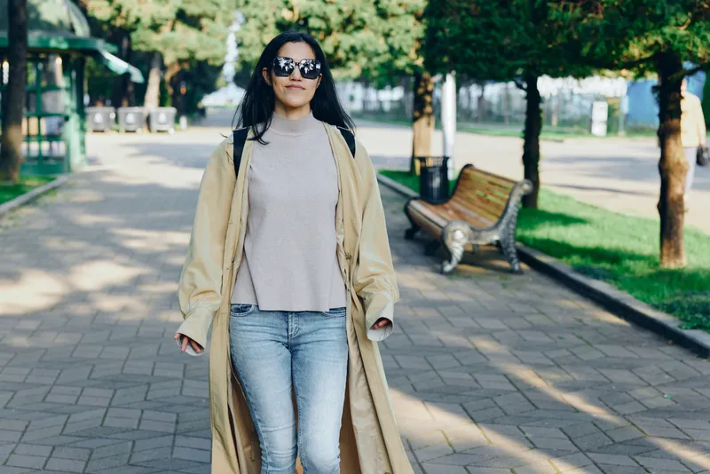 Woman wearing a long beige trench coat and gray sweater walks along a sunlit park path with sunglasses, urban style, confident stride and relaxed mood, modern fashion scene in a casual outdoor setting