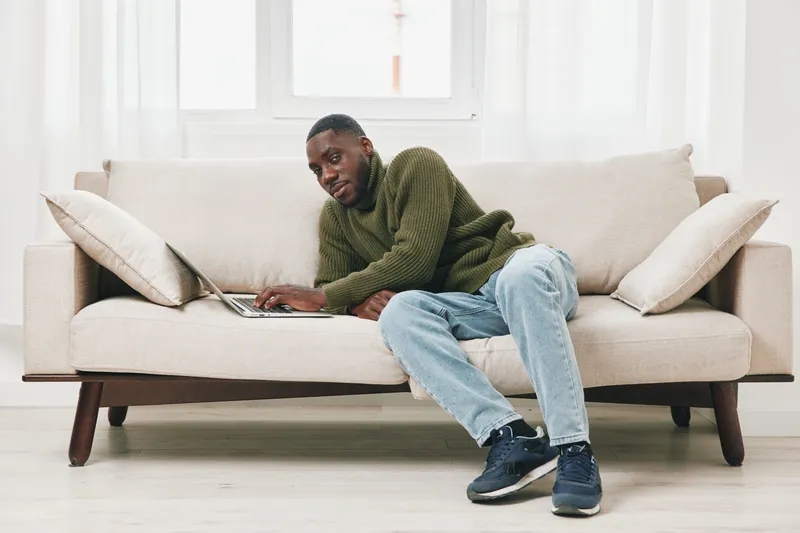 Young Black man working on a laptop at home, wearing a casual green sweater and relaxed jeans, expresses focus in a bright and cozy living room