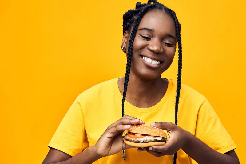 Smiling Black girl holding a delicious burger against a vibrant yellow background, showcasing joy and a casual lifestyle with a focus on healthy eating