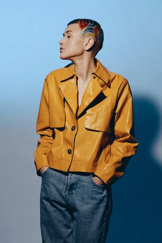 Stylish woman with short colorful hair wearing a yellow jacket and casual pants, looking sideways against a soft blue background, showcasing modern fashion and attitude.