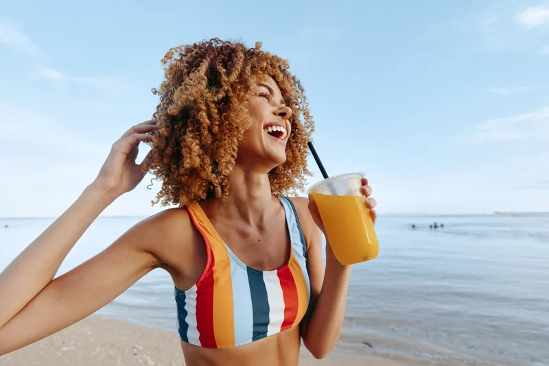 Laughing woman with curly hair on a beach, striped swimsuit, holding a yellow drink cup, summer vacation, bright daylight and calm sea horizon