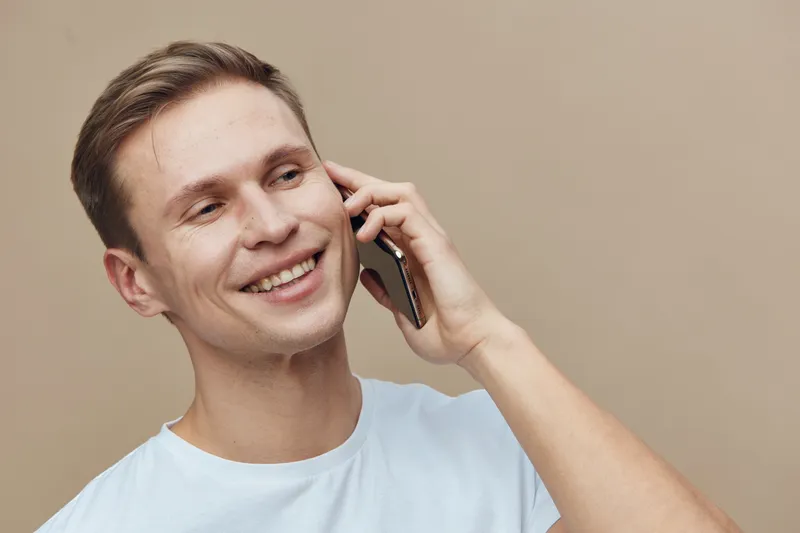 smiling young caucasian man holding smartphone talking on phone casual white t-shirt beige background digital communication concept