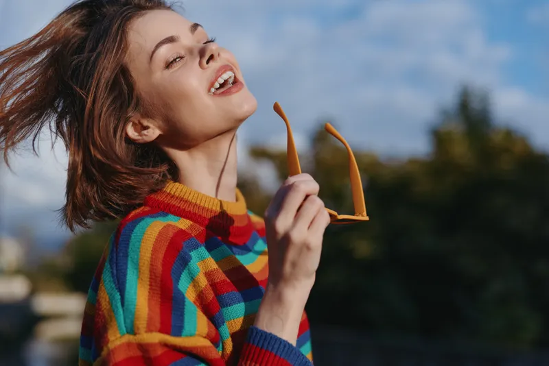 Young woman in a striped sweater holds sunglasses while smiling outdoors, mid twenties model with short brown hair and freckles, casual outfit, joyful travel lifestyle portrait.