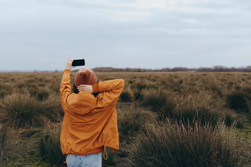 Woman lifestyle outdoors in an autumn field, wearing a mustard jacket and white scarf, arms raised as wind lifts fabric, soft daylight, distant grass, open sky, calm nature mood