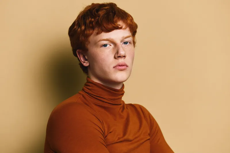 Young man with red hair and fair skin wearing brown turtleneck posing against beige background. Studio portrait with serious expression and soft lighting for fashion or modeling.