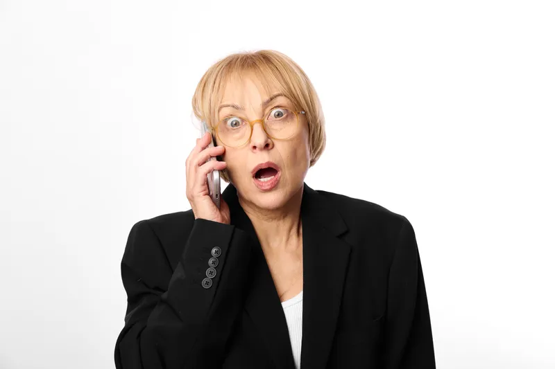 Surprised elderly woman in business suit holds phone call to ear, wide eyes, shocked expression in minimalist studio portrait. Concept for stress, communication, and crisis.