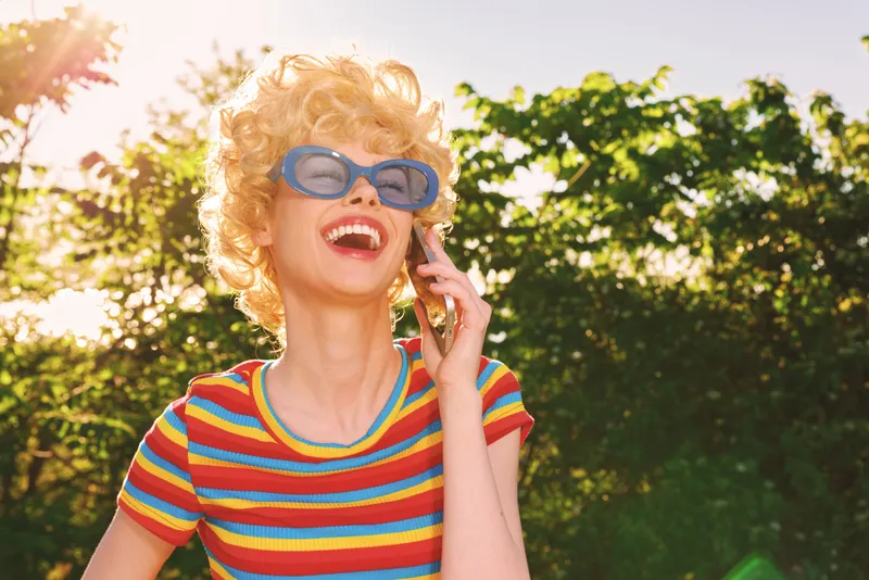 Joyful woman with curly blonde hair wearing a striped colorful shirt and oversized sunglasses talks on a mobile phone outdoors, sunny garden mood, retro lifestyle scene