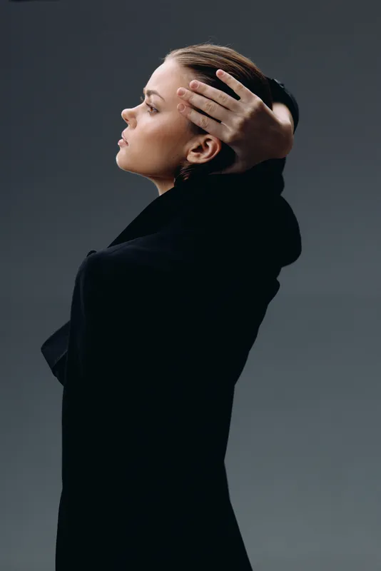 Profile of a thoughtful woman with braided hair wearing a black outfit against dark background.