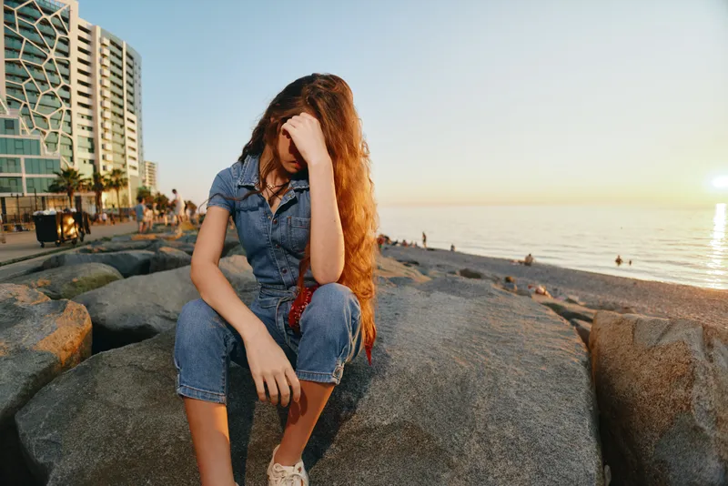 Lifestyle woman in modern boho-western denim street style sitting on rocks by beach during golden hour with soft film color lighting