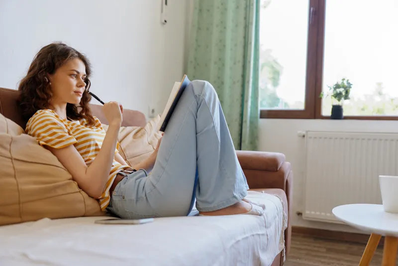 Relaxed woman with curly hair lounging on a sofa using a tablet in a bright living room, creating a cozy home atmosphere, casual lifestyle, self care and focused mood.