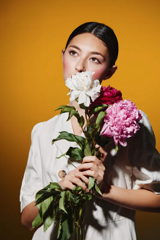 Young woman in a white dress holding white and pink flowers close to her face on a yellow background. Creative beauty and fashion concept with soft lighting and natural elegance.