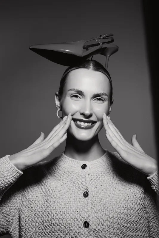Smiling white woman with light skin and dark hair balances high heel shoe on head, wearing textured sweater and hoop earrings in studio. Concept of fun and fashion.
