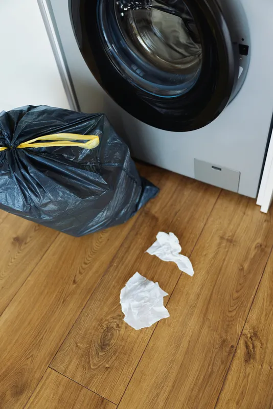 Washing machine and trash bag on a wooden floor with crumpled tissues scattered around, laundry room mess and household waste beside the appliance in a small domestic utility corner.