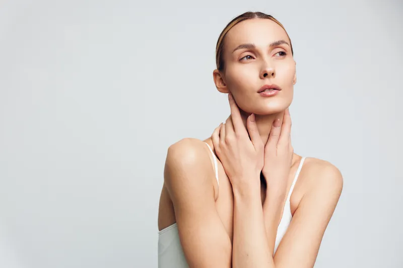 Young woman with clear skin posing with hands gently touching neck, minimal makeup, soft lighting, neutral background, calm expression, beauty concept, skincare routine.