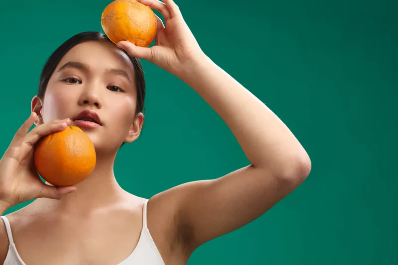 Asian woman poses with oranges in a vibrant studio, holding fruit near her face and smiling. The bright setup creates a fresh, playful and healthy mood.