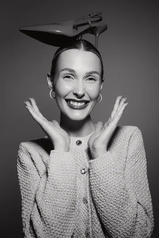 Smiling white woman with fair skin holds hands near face with high heel shoe balanced on head in studio.