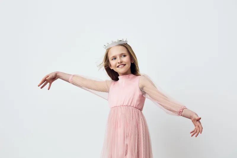 Joyful girl in a pink dress and crown, exuding happiness against a light gray background, perfect for children s fashion and celebrations