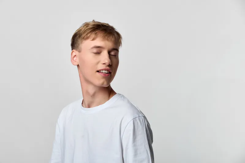 Calm, relaxed youth wearing a white tee with closed eyes, soft smile, against a neutral background, conveying serenity, introspection, and casual comfort.
