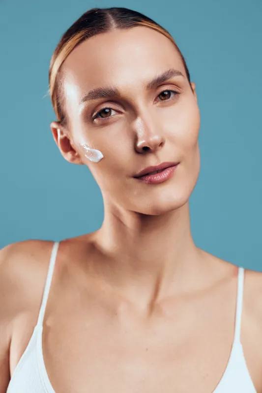 Young woman with clear skin applying moisturizing cream on cheek, natural beauty concept, soft lighting, smooth complexion, blue background, skincare routine